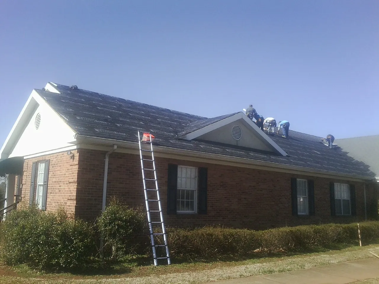 Crew tearing shingles off a brick ranch roof with a ladder set — Metal Roof Repair work by our crew in Holley