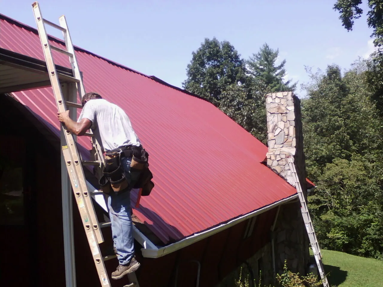 Roofer on a ladder beside a red metal roof and stone chimney — Soffit & Fascia Repair by Roofing Brothers in Holley